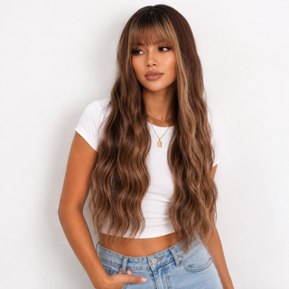 A woman modeling a long, wavy wig with dark brown roots and warm caramel-brown highlights, styled with straight-cut bangs. She is wearing a white crop top and light blue jeans against a plain white background.